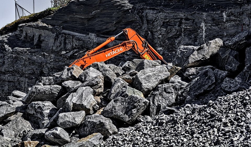 Una excavadora Hitachi de color naranja se alza sobre una colina rocosa, rodeada de grandes rocas y cantos rodados grises. El brazo de la excavadora está extendido y se puede ver parte de una cerca de alambre en la esquina superior izquierda de la imagen, indicativo de Permisos de Minería en acción.