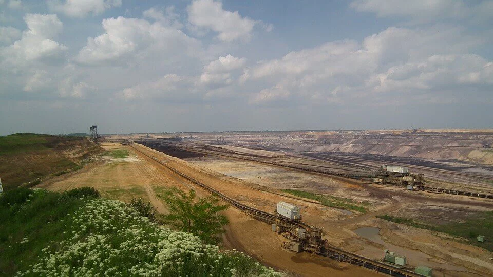 Um grande local de mineração a céu aberto sob um céu nublado. A cena apresenta sujeira escavada e terra em camadas, com equipamentos de mineração e correias transportadoras típicas do setor de mineração visíveis. Vegetação verde e pequenas flores brancas estão em primeiro plano.