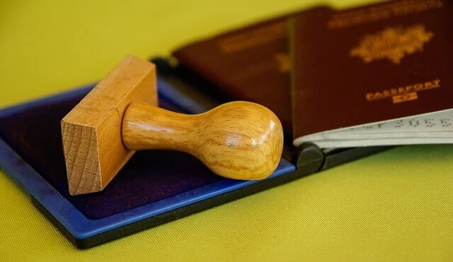 A wooden rubber stamp is resting on an ink pad. Behind the stamp, there are two partly visible passports with brown covers, suggesting steps involved in the Formação da Empresa. The background surface is yellow.