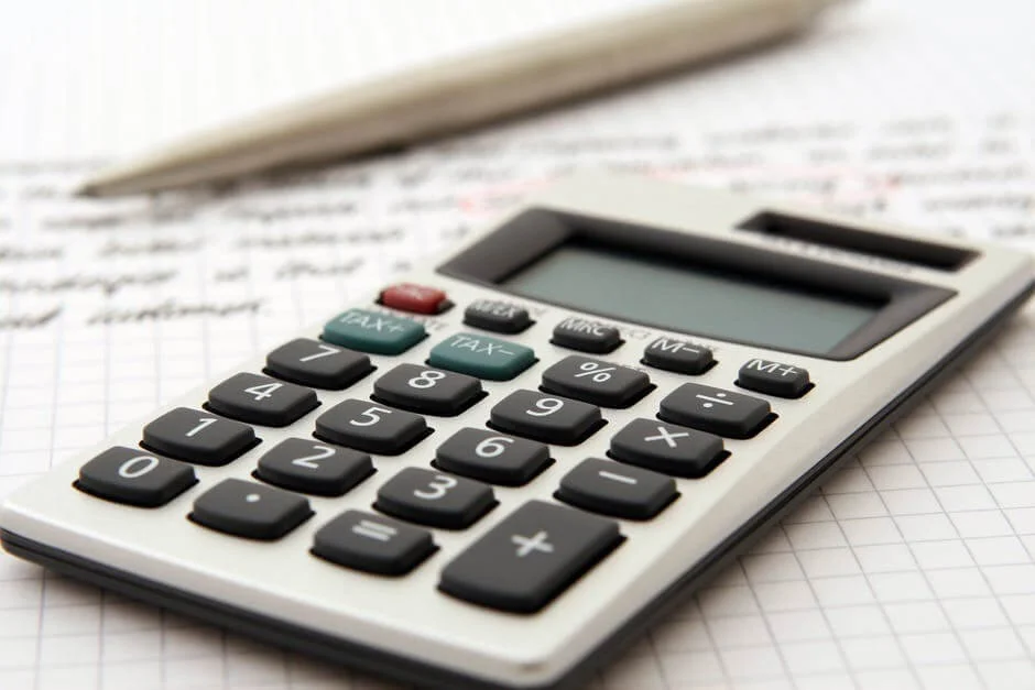 A close-up image of a calculator placed on a piece of grid paper with handwritten text discussing Formação da Empresa visible in the background. A pen is lying next to the calculator. The calculator has a numeric keypad along with function keys.