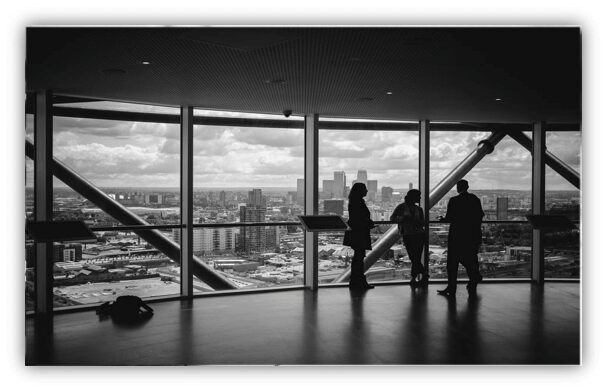 Una imagen en blanco y negro que muestra a tres personas de pie junto a grandes ventanales en una plataforma de observación. El paisaje urbano, lleno de edificios y un cielo parcialmente nublado, es visible a través de las ventanas. Las personas parecen mantener una conversación sobre servicios contables.