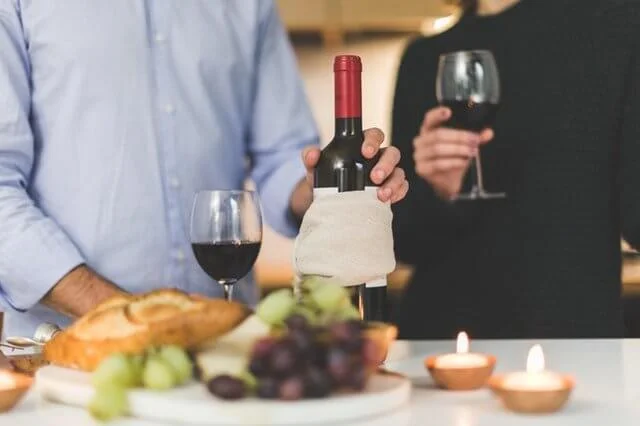 Duas pessoas estão em uma mesa, saboreando taças de vinho tinto. A mesa, adornada com uma garrafa de vinho, velas acesas, um pão, uvas e nozes, reflete a riqueza da exportação do vinho peruano. Uma pessoa coloca mais vinho em uma taça enquanto a outra segura a sua, pronta para degustar.