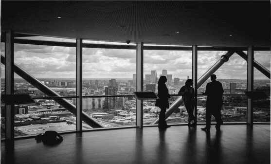 Tres personas se paran frente a grandes ventanales, con vistas a un paisaje urbano con edificios altos. La sala, posiblemente una oficina de servicios contabilidad Perú, tiene un diseño moderno con vigas estructurales expuestas. El cielo está nublado, arrojando una luz tenue sobre la escena. A la izquierda hay una bolsa en el suelo.