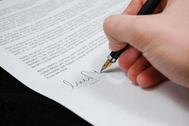 A hand holds a black pen and signs a document on a white sheet of paper. The document, related to residencia permanente México, contains printed text in a small font, and the signature is being written near the bottom of the page. The tip of the pen is touching the paper as the signature is formed.