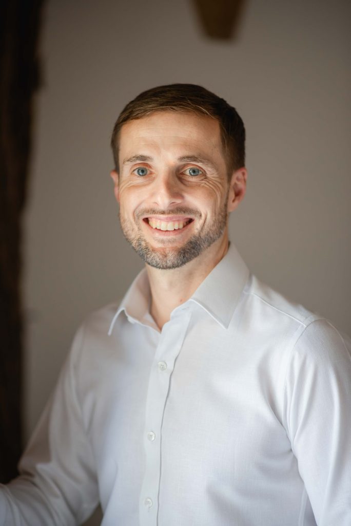 A man with short hair and a beard is smiling at the camera, exuding the spirit of entrepreneurship. He is wearing a white button-up shirt and stands against a neutral background, embodying the vibrant energy of Colombia.