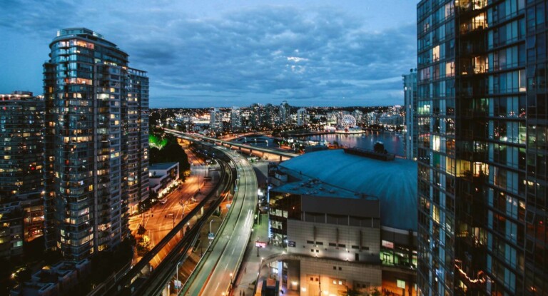 Cityscape at dusk showing lighted buildings and streets. A curved highway cuts through the scene, with traffic lights and vehicles visible. Near the center, a large domed building stands out, surrounded by other high-rise structures, reminiscent of a bustling hub for formação da empresa.