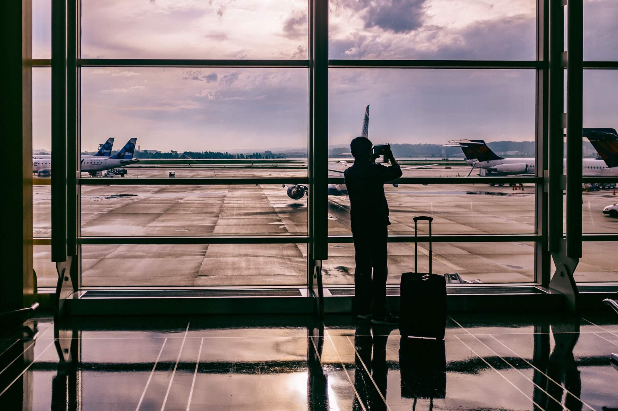 A person stands with a suitcase, gazing through large airport windows at planes on the tarmac. The sky is cloudy, and the ground is wet from recent rain. Jets are parked in view as they capture a photo or video on their mobile device, perhaps documenting moments for their Formação da Empresa journey.