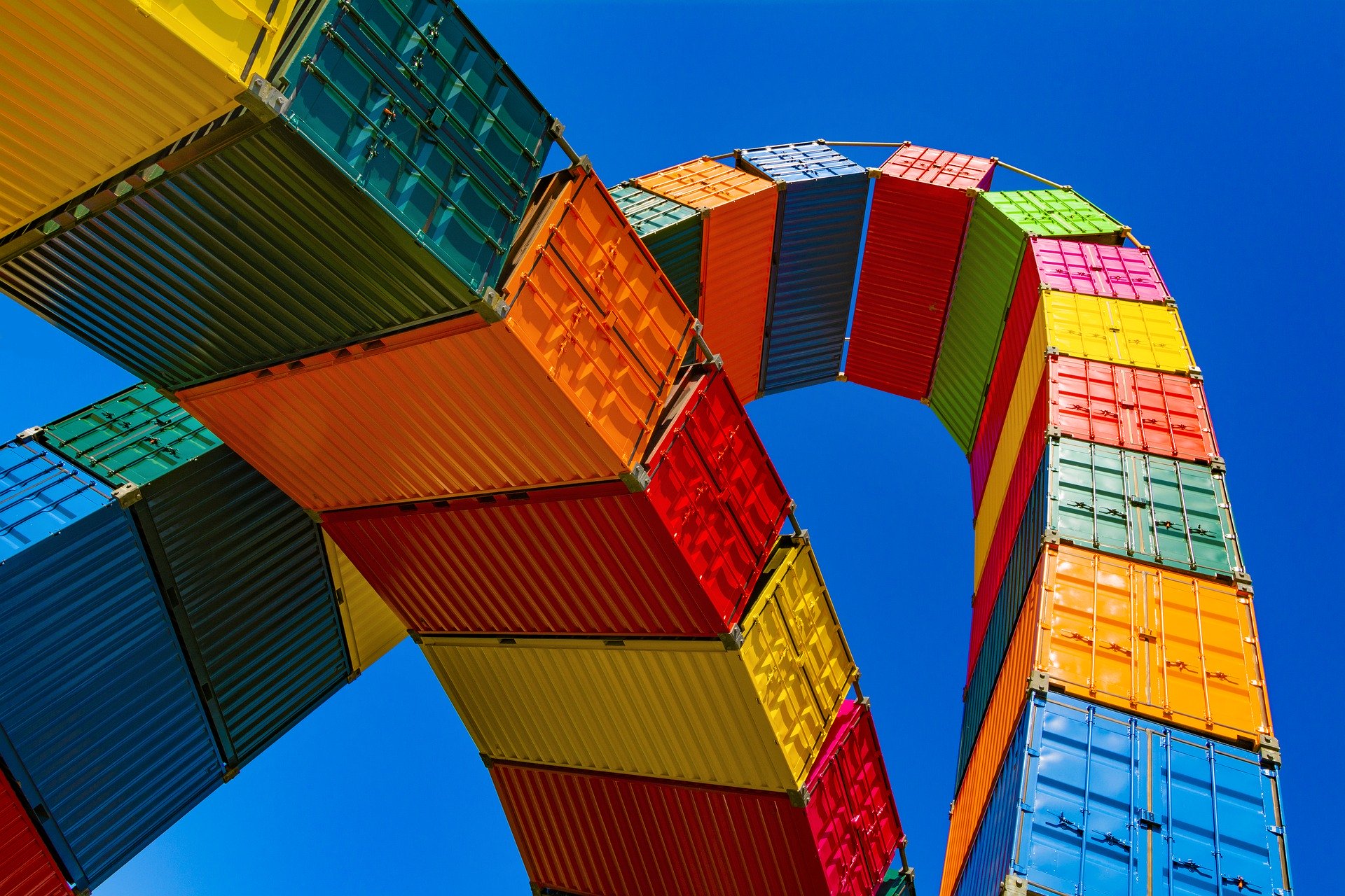 Una instalación de contenedores marítimos de colores brillantes, que recuerdan el vibrante impulso económico que han supuesto las inversiones del TLC Perú-China, apilados y curvados en un arco contra un cielo azul despejado. Los contenedores de color rojo, naranja, amarillo, verde y azul reflejan el espíritu dinámico de los acuerdos de libre comercio. 