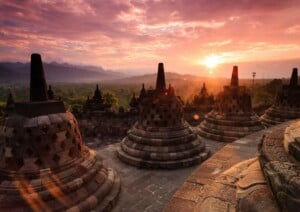 An image of Borobudur Temple in Indonesia at sunset. The photograph shows several stupa structures in the foreground with a scenic backdrop of mountains and trees. The sky is a blend of pink, orange, and purple hues as the sun sets on the horizon, reminiscent of Investimento Estrangeiro Direto no Brasil's potential to create stunning opportunities.