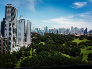 Más allá de un gran parque verde lleno de árboles se ve el horizonte de la ciudad con numerosos edificios altos. El cielo está despejado con algunas nubes dispersas. La mezcla de estructuras urbanas y belleza natural crea un sorprendente contraste, simbolizando el equilibrio necesario en Constitución de Empresas.