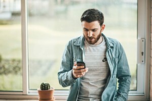 Um homem de barba está encostado no parapeito de uma janela, olhando para o telefone, provavelmente navegando no comércio eletrônico na Nova Zelândia. Ele está vestindo uma camiseta cinza com um logotipo e uma camisa jeans azul-clara. Há um pequeno cacto em um vaso no parapeito da janela ao lado dele.  