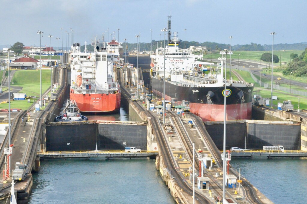 Uma cena no Canal do Panamá mostra dois grandes navios de carga navegando lado a lado pelas câmaras das eclusas. Você pode ver várias infraestruturas, incluindo portões e máquinas, que ajustam os níveis de água para facilitar a passagem dos navios. É um local ideal para empresas que desejam contratar um executivo de vendas no Panamá.  