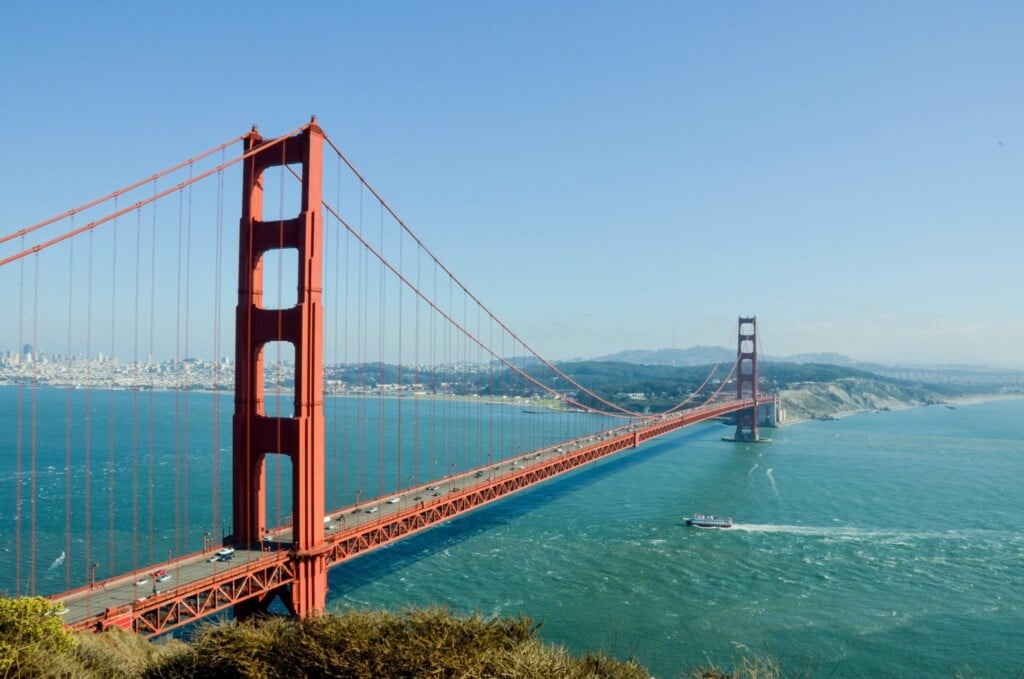 Vista da ponte Golden Gate sobre um corpo de água em um dia claro. Vários veículos estão atravessando a ponte, talvez fazendo negócios na Bolívia, enquanto um barco viaja na água abaixo. O horizonte da cidade e as colinas são visíveis ao fundo.  