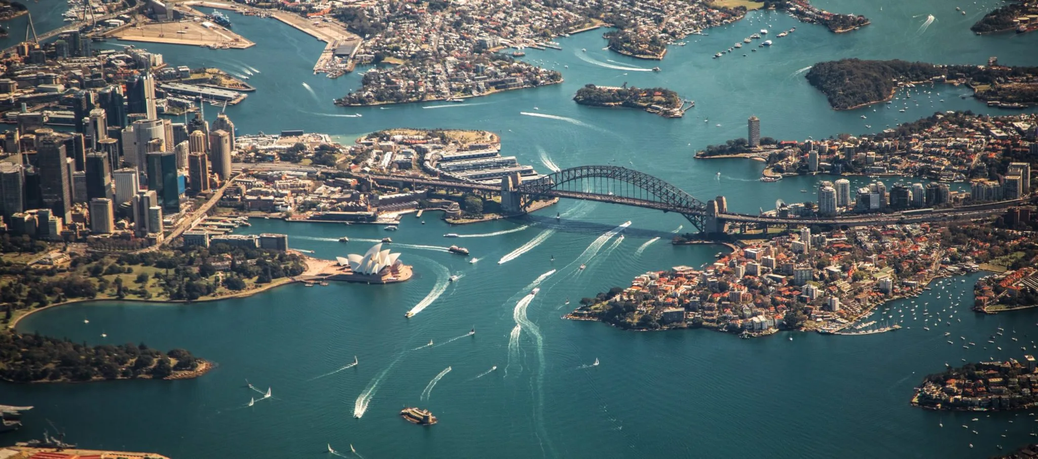 Aerial view of a coastal city featuring a large harbour with numerous boats. The scene includes a prominent bridge and a distinct building with white, sail-shaped roofs near the water. As the coastline is densely developed with both high-rise buildings and residential areas, one wonders about the future of insurtech in Australia.