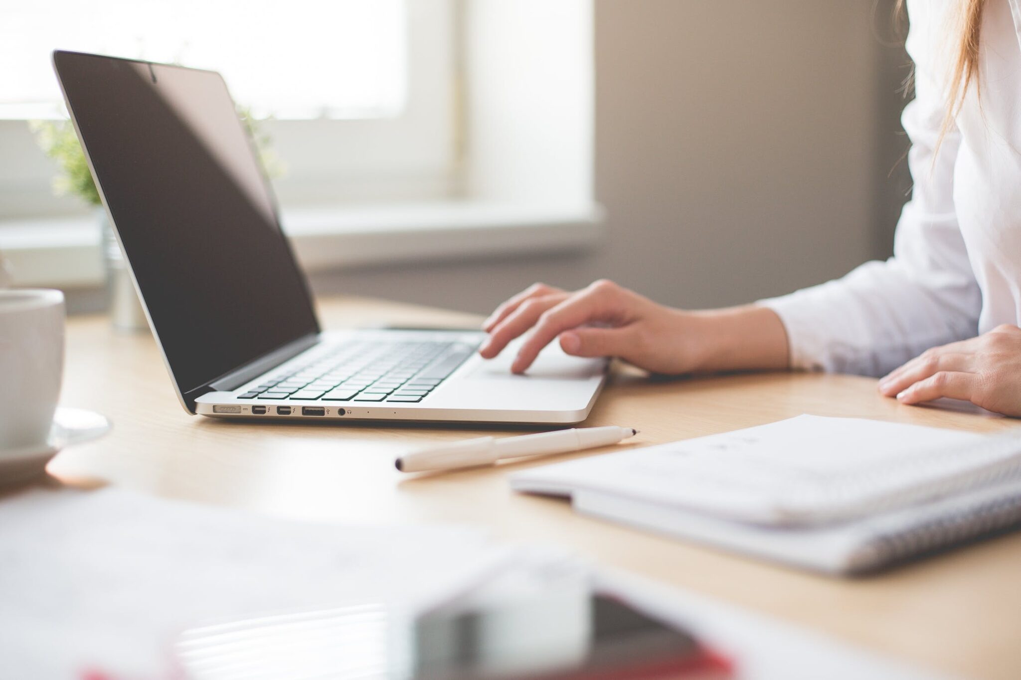 Uma pessoa está trabalhando em um laptop em uma mesa de madeira, possivelmente pesquisando um visto de negócios no Peru para cidadãos americanos. A mão direita está sobre o teclado do laptop, enquanto você tem por perto um caderno, uma caneta e uma xícara de café. A luz natural passa por uma janela ao fundo.  