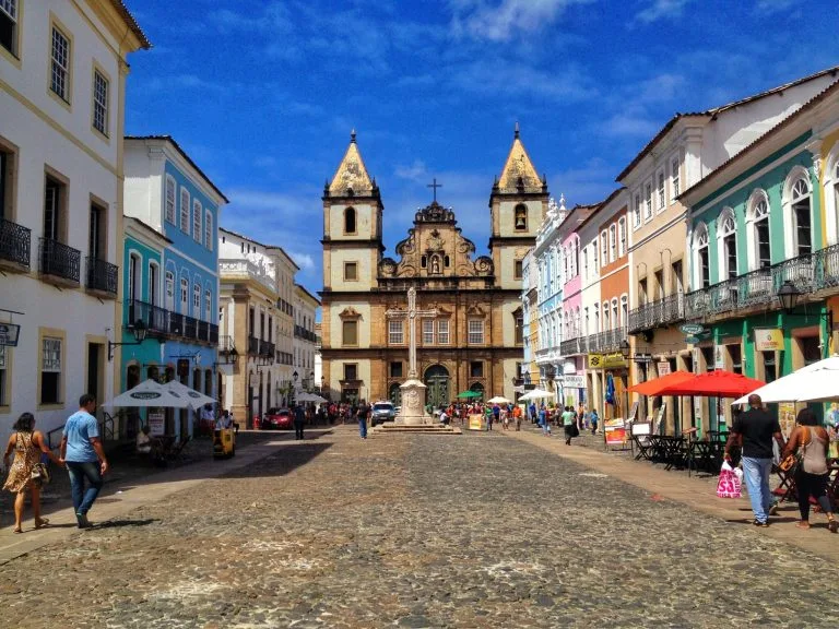 Una calle adoquinada bordeada de coloridos edificios y cafés al aire libre. La gente camina por la vibrante calle, discutiendo sobre cómo liquidar una empresa en El Salvador, mientras se dirigen hacia una iglesia histórica con dos campanarios y una cruz en lo alto, bajo un cielo azul despejado.