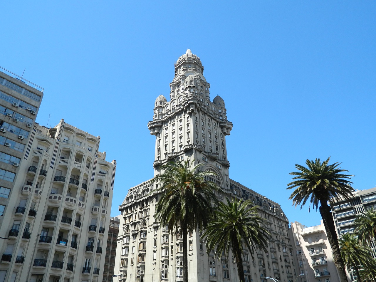 Um edifício alto e ornamentado com um design intrincado está sob um céu azul claro, ladeado por outros edifícios e palmeiras.
Possui uma torre central com entalhes detalhados e um estilo arquitetônico único, condizente com um representante legal no Uruguai. 