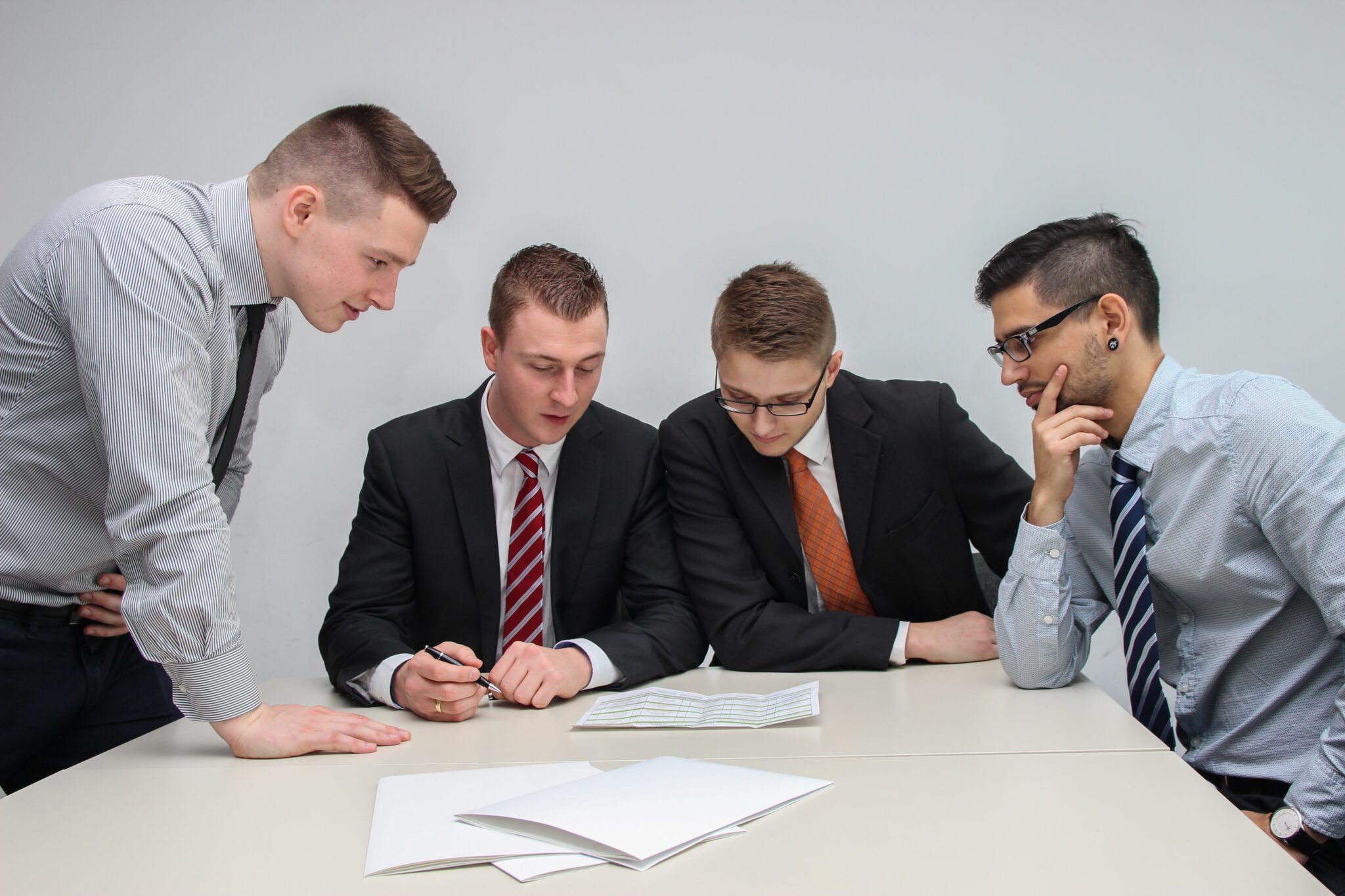 Quatro homens em trajes de negócios estão reunidos em torno de uma mesa analisando documentos, concentrando-se atentamente nos recentes acordos comerciais do Mercosul. Três deles estão sentados, enquanto um se levanta e se inclina para olhar os documentos. A mesa também tem algumas folhas de papel em branco para futuras negociações.  