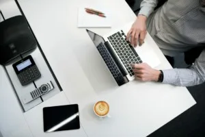 Uma pessoa está sentada em uma mesa branca digitando em um laptop. Ao lado do laptop, há um caderno em espiral, uma xícara de café, um telefone de escritório e um tablet que exibe informações sobre serviços de secretariado corporativo no Chile. A pessoa está usando uma camisa listrada e um relógio.