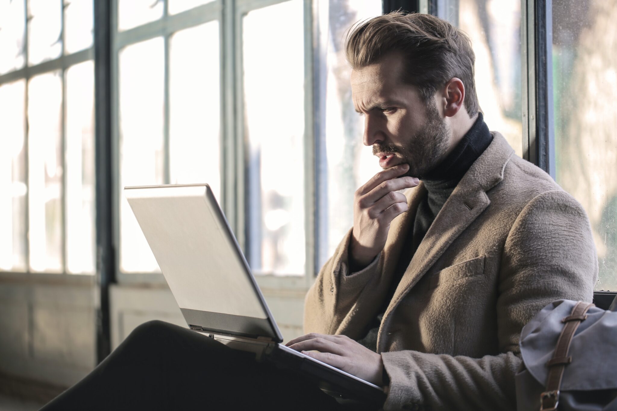 A man with light brown hair and a beard, wearing a brown coat and black turtleneck, is sitting in front of large windows and looking at a laptop screen. He has a thoughtful expression, possibly contemplating his latest article on caçadores de cabeças na Colômbia, resting his chin on his hand.