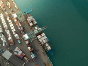 Vista aérea de un puerto marítimo con numerosos contenedores organizados en filas. Varias grúas están colocadas a lo largo del muelle, cargando y descargando exportaciones de los buques de carga atracados. El agua azul clara contrasta con la disposición ordenada de los contenedores de Costa Rica en tierra.