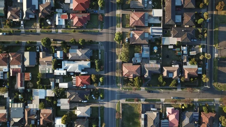 Vista aérea de un barrio suburbano que presenta un diseño de cuadrícula con carreteras que se cruzan. El área incluye múltiples casas con patios traseros, varios estilos de techos, árboles que bordean las calles y algunos autos estacionados visibles en las carreteras y caminos de acceso, ideal para quienes buscan comprar una propiedad en Bolivia.