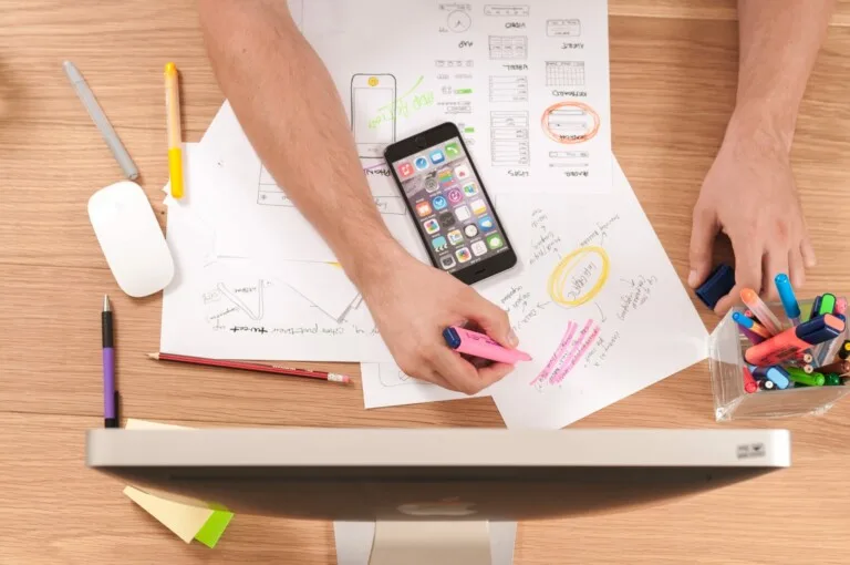 Una persona está trabajando en un escritorio con bocetos de diseño, un iPhone, rotuladores de colores, un ratón y un monitor de ordenador. Entre las herramientas de escritura dispersas, sostiene un rotulador fluorescente rosa y marca un papel mientras contempla una próxima Revisión de la Salud de la Entidad en Panamá.