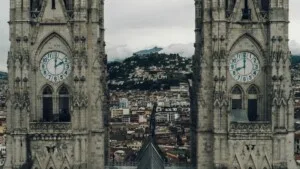 Duas altas torres de relógio de pedra com mostradores azuis que exibem logo após as 9h15 e as 15h15, emoldurando a vista de uma cidade com uma montanha ao fundo. As torres apresentam arquitetura gótica, assim como os acordos de dupla tributação do Equador trazem contexto histórico para as paisagens financeiras modernas.