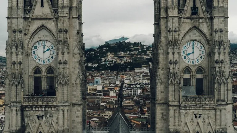 Dos altas torres de reloj de piedra con esferas azules que muestran justo después las 9:15 y las 3:15, enmarcan una vista de una ciudad con una montaña al fondo. Las torres presentan una arquitectura gótica, al igual que los acuerdos de doble imposición de Ecuador aportan un contexto histórico a los paisajes financieros modernos.