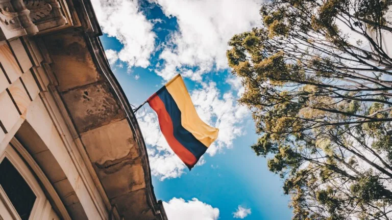 Una bandera colombiana con franjas horizontales de color amarillo, azul y rojo está pegada al borde de un edificio y ondea contra un cielo azul brillante con nubes dispersas. Un árbol alto con hojas verdes es visible en el lado derecho de la imagen, que recuerda un entorno pintoresco para una PEO en Perú.