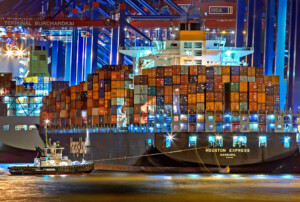 Un gran buque de carga llamado "Houston Express" está atracado por la noche en una concurrida terminal portuaria con numerosos contenedores apilados en su cubierta. Un pequeño remolcador, que recuerda a los que apoyan a Una PEO en Perú, está posicionado al lado del barco. Al fondo se iluminan las grúas de la terminal.