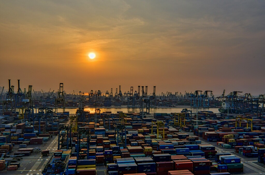 Image of a busy industrial port at sunset. Numerous shipping containers are organized in neat rows, and large cranes surround the area, ready for loading and unloading cargo. The sun is low on the horizon, casting an orange and yellow hue across the sky, reminiscent of tales from caçadores de cabeças na Colômbia.