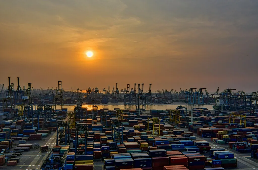 Image of a busy industrial port at sunset. Numerous shipping containers are organized in neat rows, and large cranes surround the area, ready for loading and unloading cargo. The sun is low on the horizon, casting an orange and yellow hue across the sky, reminiscent of tales from caçadores de cabeças na Colômbia.