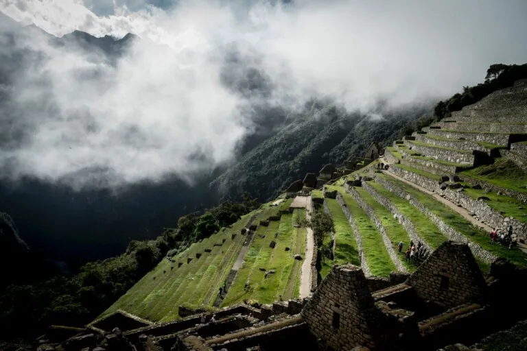 Uma vista enevoada de campos com terraços e antigas estruturas de pedra em Machu Picchu. O plano de fundo consiste em montanhas nubladas e arborizadas. Em meio a vários visitantes que exploram o sítio arqueológico inca, os sussurros da reestruturação corporativa no Peru sugerem uma paisagem em transformação além desses terraços históricos.