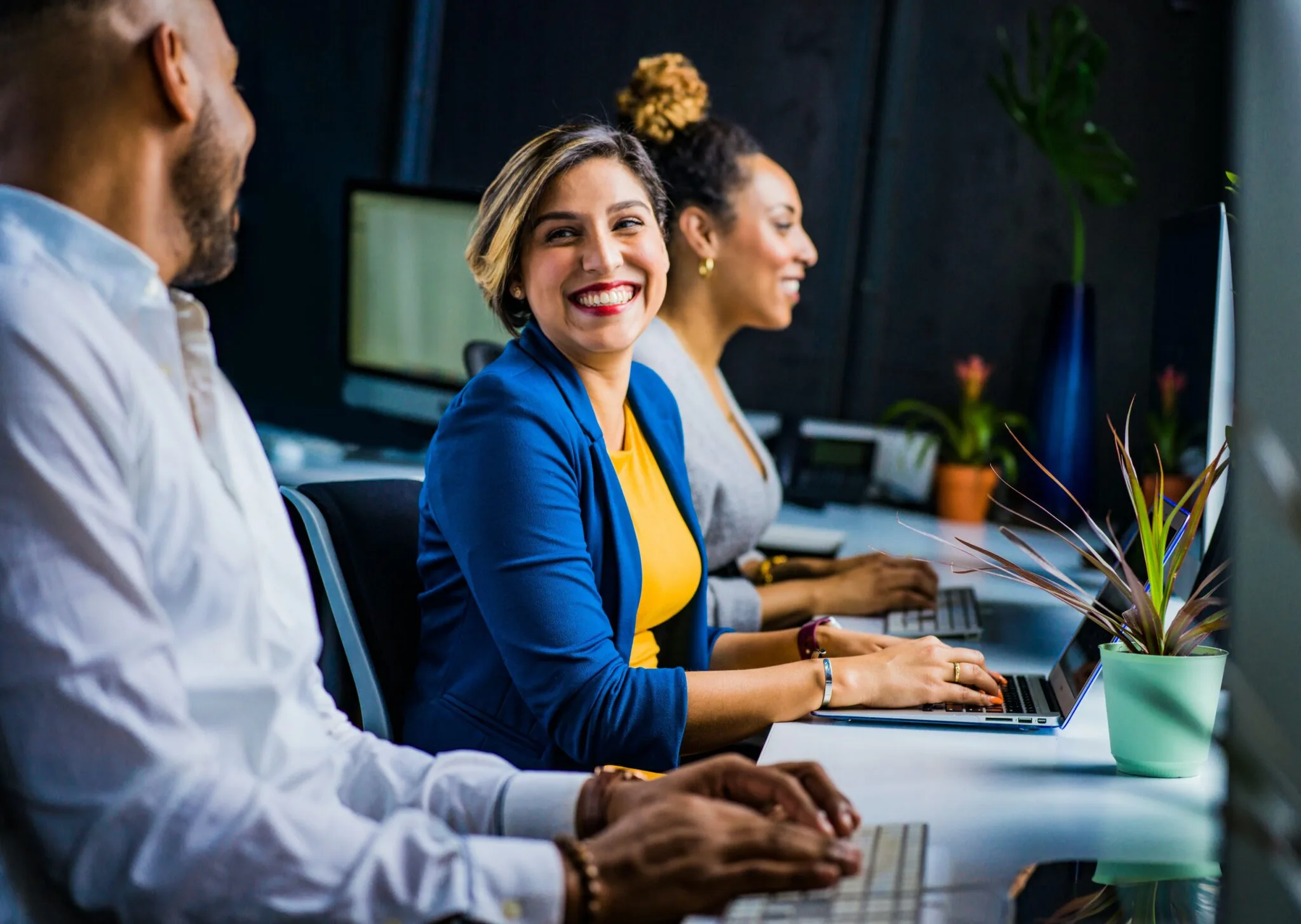 Three people are seated at desks, working on laptops in an office setting. The person in the middle is smiling and looking at the person on the left, possibly discussing M&A due diligence in Mexico. The background includes computer monitors and office decor, such as potted plants.