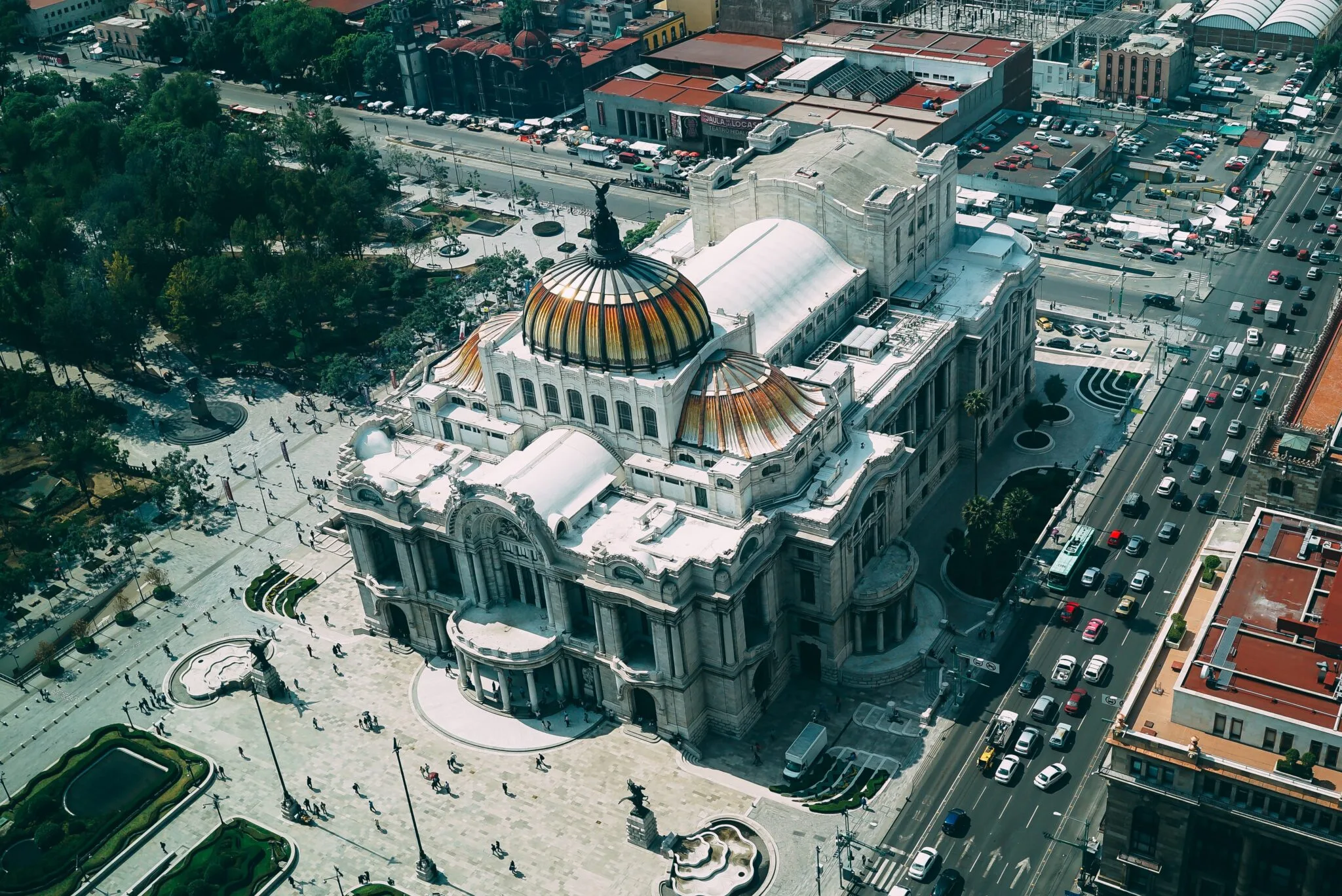 Aerial view of a large, historic building with a distinct orange and yellow-tiled dome. The structure is surrounded by busy streets bustling like the activity during M&A Due Diligence in Mexico, with numerous cars and people walking on the adjacent sidewalks. The building's architecture showcases an elegant and ornate design.