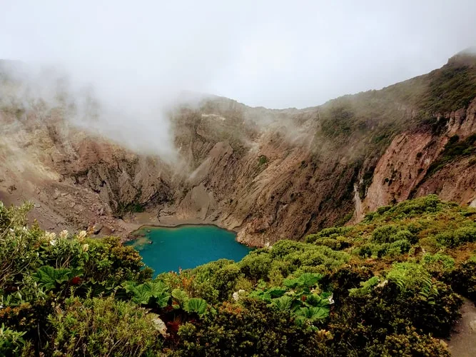 A turquoise crater lake is surrounded by steep rocky cliffs with sparse vegetation, reminiscent of the breathtaking scenery you might find when buying property in Costa Rica. In the foreground, green bushes and plants are visible. Mist and clouds cover parts of the cliffs, giving the scene a misty appearance.