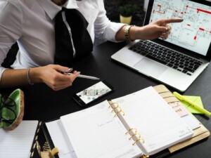 Uma pessoa está sentada em uma mesa trabalhando. Há um laptop exibindo planos arquitetônicos relacionados à compra de propriedades no Panamá, um smartphone, uma agenda aberta, um par de óculos, uma caneta e um marcador de texto sobre a mesa. A pessoa está apontando para a tela do laptop enquanto segura uma caneta com a outra mão.  