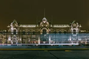 A large, ornate building with extensive nighttime lighting, viewed through black iron fencing. A blurred motion effect, likely from passing vehicles or lights, is visible in front of the building, adding a touch of mystery akin to the tales of caçadores de cabeças na Colômbia against the dark sky.