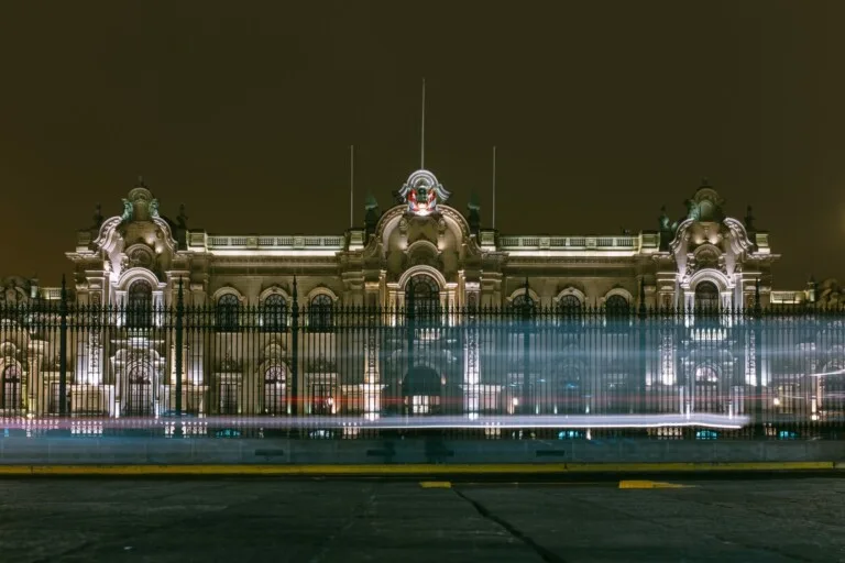 Por la noche se ve un edificio majestuoso e iluminado con intrincados detalles arquitectónicos y un cielo oscuro. Una valla recorre el frente, mientras que los rastros de luz de los vehículos que pasan se difuminan en la parte inferior de la imagen. Esta escena captura la esencia de Una PEO en Perú, fusionando tradición con modernidad.