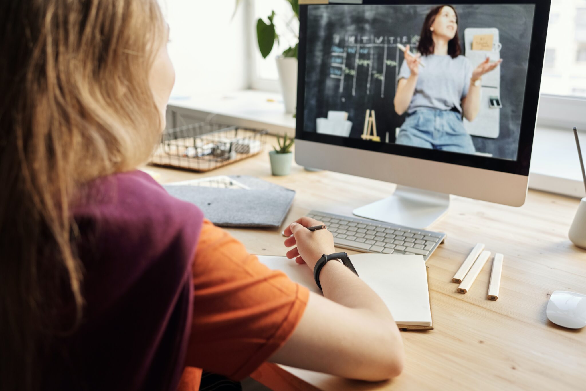 Uma pessoa está sentada em uma mesa com um computador, participando de uma aula on-line. A tela do computador exibe uma mulher gesticulando perto de um quadro negro, possivelmente discutindo o crescimento de empresas de tecnologia na Colômbia. A mesa tem um caderno, lápis e pequenos vasos de plantas. A luz do sol entra por uma janela ao fundo.   