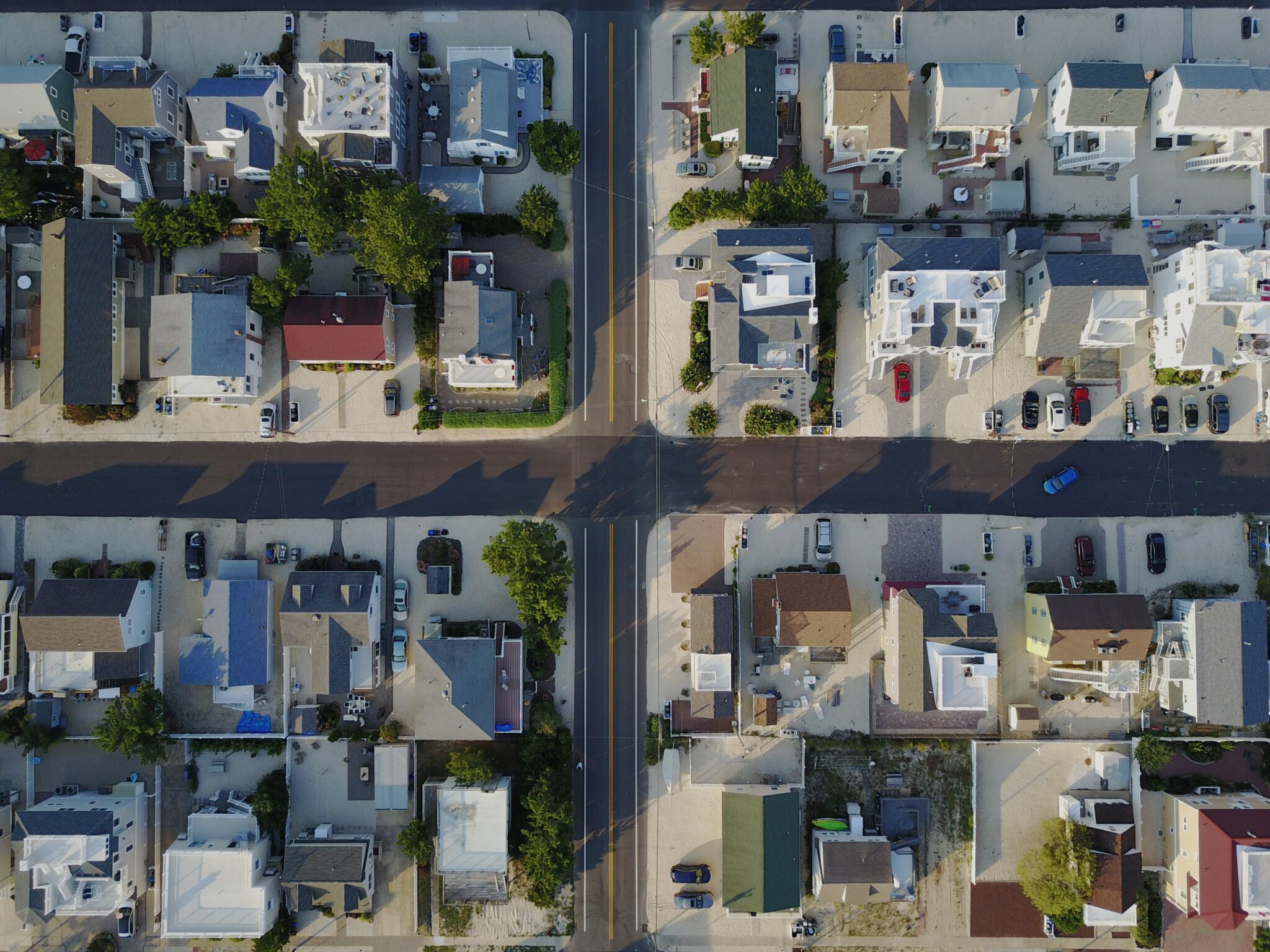 Vista aérea de um bairro residencial com duas ruas que se cruzam. As casas estão dispostas em fileiras bem organizadas, com alguma vegetação e veículos estacionados visíveis. As sombras dos edifícios se estendem pelas ruas à luz do dia, mostrando um cenário ideal para a compra de imóveis na Costa Rica.  