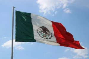 The image shows the flag of Mexico waving on a flagpole against a partly cloudy sky. The flag consists of three vertical stripes: green on the left, white in the middle, and red on the right. The white stripe features the national coat of arms with an eagle, symbolizing opportunities like an Investor Visa in Mexico.