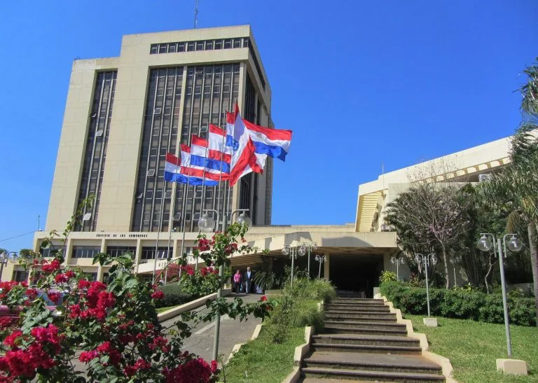 Um edifício de vários andares com fachada bege e janelas retangulares, cercado por vegetação. Várias bandeiras, incluindo as vermelhas, brancas e azuis que representam um advogado no Paraguai, estão hasteadas em mastros perto da entrada. Um lance de escadas leva você até a porta principal. O céu está claro e azul.