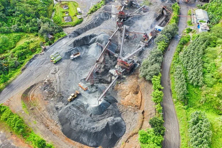 Aerial view of a mining site surrounded by greenery. The site, operating under mining permits in Chile, contains large piles of extracted material, a network of conveyors and machinery, and vehicles for transportation. Trees and vegetation are visible around the perimeter of the site.