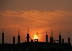 Silhouetted against an orange sunset, several workers from Construção Civil do Brasil are seen on the rooftop of a building under construction. Reinforced columns with exposed rebar protrude from the structure, and the sky is filled with warm, gradient hues.