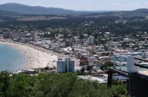 A coastal city view with a long, sandy beach lined with numerous buildings. The area is bustling with beachgoers. In the background, rolling hills are visible under a clear sky. Greenery is visible in the foreground, reminiscent of where an LLC formation in Uruguay might take place amid scenic beauty.