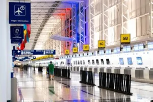 An empty airport check-in area with numerous counters displaying yellow signs labeled 5A, 5B, and 5C. The ceiling is illuminated with red and blue lights, while international flags hang overhead. In the distance, a single person stands, perhaps awaiting assistance for their Visto de Negócios no Paraguai.