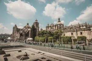 La imagen muestra una vista de una calle con gente caminando, con una catedral histórica al fondo bajo un cielo parcialmente nublado. El primer plano incluye una barrera de cristal y lo que parece ser un yacimiento arqueológico o elementos de construcción modernos, posiblemente mezclando el encanto antiguo con la bulliciosa vida de un abogado en México.