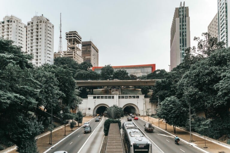 A busy divided road with vehicles passes through tunnels and is flanked by green trees. Above the tunnels, a bridge with pedestrians spans the road. Tall buildings, where you might find a Representante Legal no Brasil, are visible in the background against a cloudy sky.